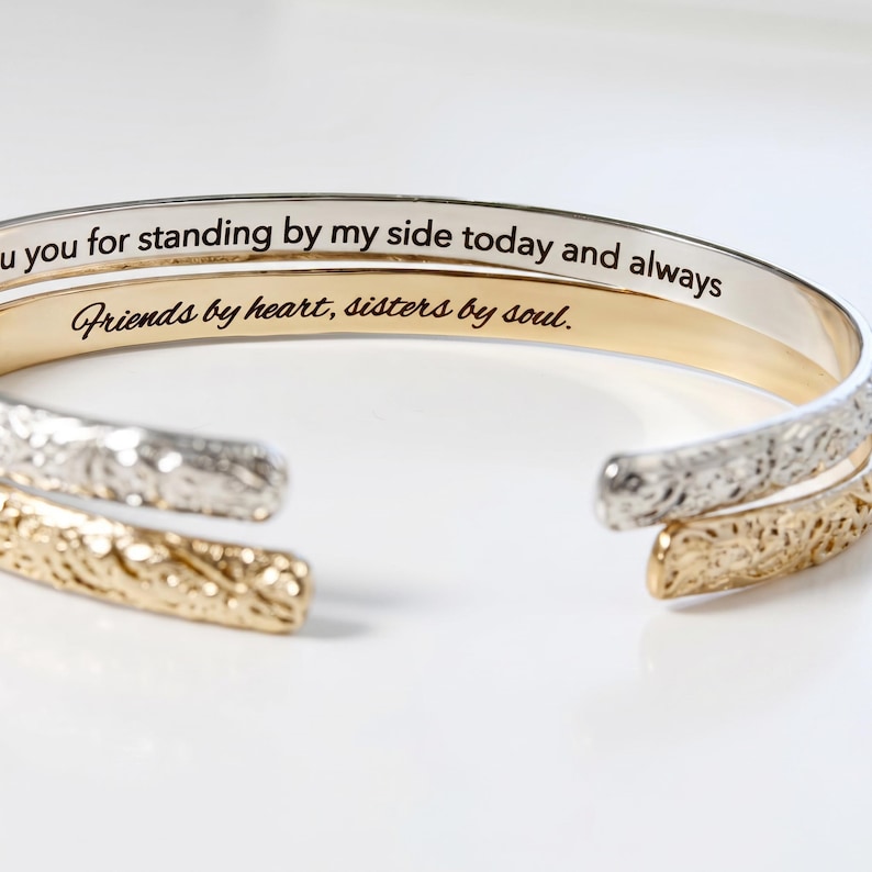 Two open cuff bracelets, one gold and one silver, with engraved text on the inside that reads: "Thank you for standing by my side today and always" and "Friends by heart, sisters by soul."