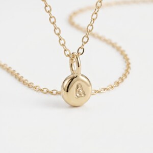 May include: A gold-colored necklace with a small, round pendant. The pendant features the letter "A" in a simple, elegant font. The chain is delicate and appears to be made of small, linked segments. The background is a soft, neutral color.