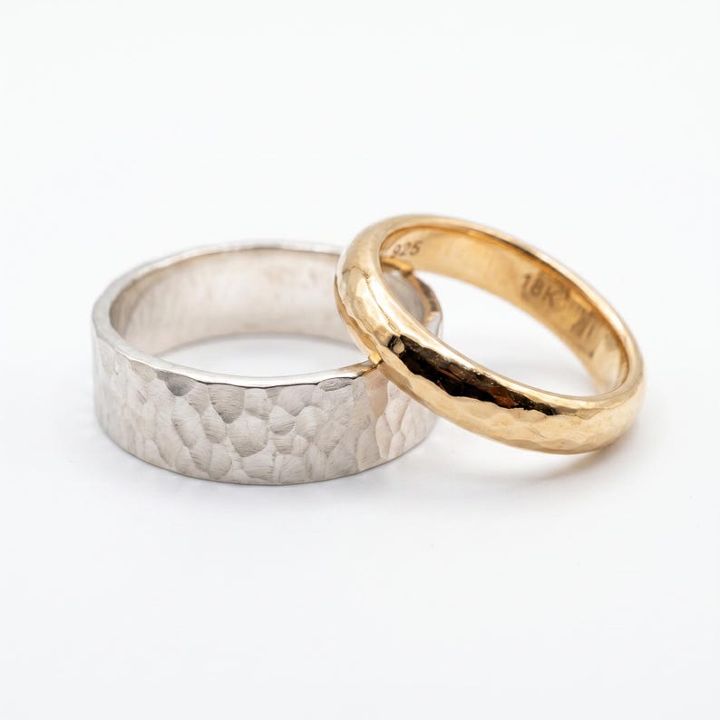 A wide, hammered silver band ring and a narrower, hammered gold band ring are stacked next to each other on a white surface.