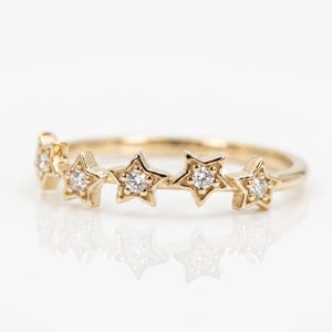 A gold ring with five diamond-studded stars across the top, reflecting on a white surface.