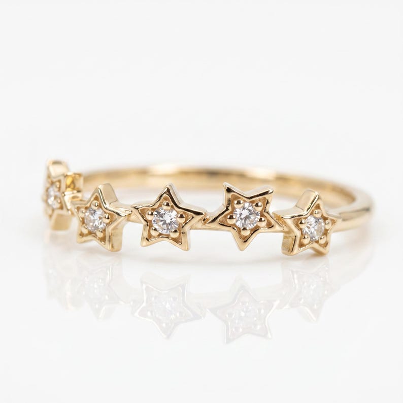 A gold ring with five diamond-studded stars across the top, reflecting on a white surface.