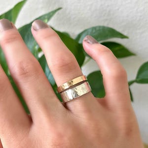 A wide, hammered silver band ring and a narrower, hammered gold band ring are stacked on a finger, with green leaves in the blurred background.