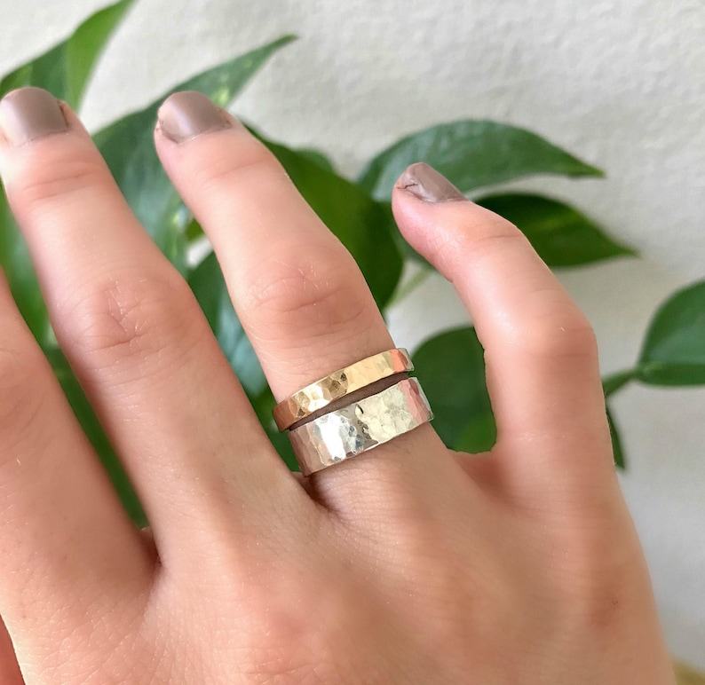 A wide, hammered silver band ring and a narrower, hammered gold band ring are stacked on a finger, with green leaves in the blurred background.