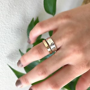 A wide, hammered silver band ring and a narrower, hammered gold band ring are stacked on a finger, with green leaves in the blurred background.