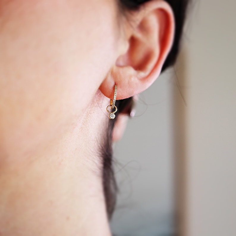 A gold hoop earring with a small diamond dangling from it. The earring is worn in the left ear of a person with dark hair.