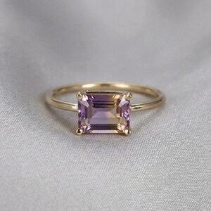 An emerald-cut ametrine ring with a thin yellow gold band rests on a grey fabric background.
