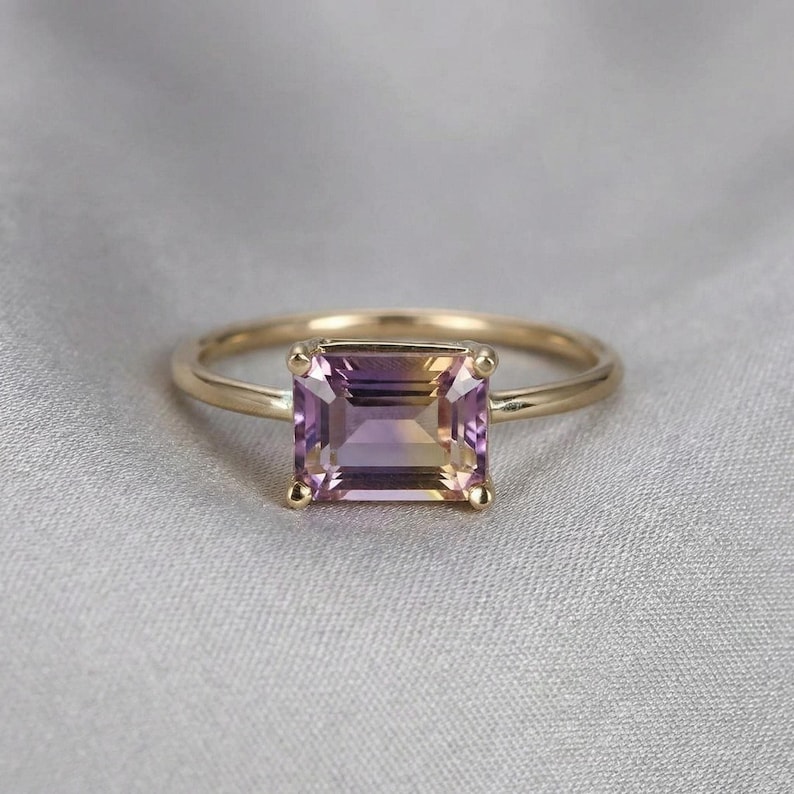 An emerald-cut ametrine ring with a thin yellow gold band rests on a grey fabric background.