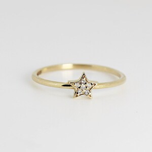 A thin gold ring with a small star shape on top, encrusted with tiny diamonds, is centered on a white background.