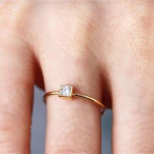A macro shot of a finger wearing a thin gold band set with a single princess cut square diamond.