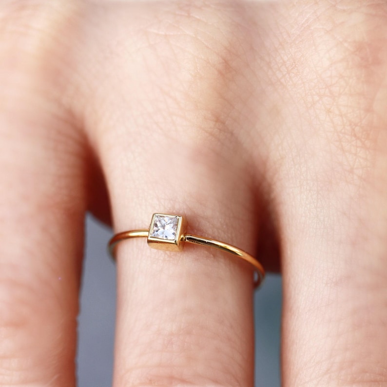 A macro shot of a finger wearing a thin gold band set with a single princess cut square diamond.