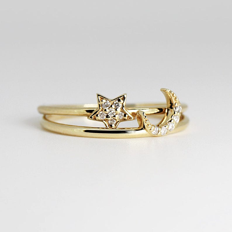 A gold ring with two bands features a diamond-studded star on one band and a diamond-studded crescent moon on the other, on a white background.