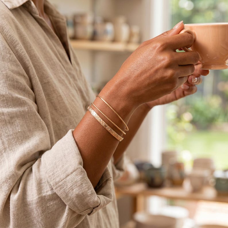 A person's arm wearing three gold bracelets: one engraved with a floral pattern, and two plain, thin bangles, holding a light brown mug.