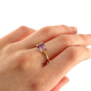 An angled view of a hand wearing an emerald-cut purple stone ring with a slim gold band.
