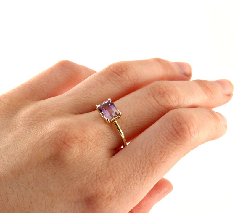 An angled view of a hand wearing an emerald-cut purple stone ring with a slim gold band.