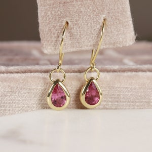 May include: A pair of gold teardrop earrings with pink gemstones. The earrings are hanging from a velvet box.