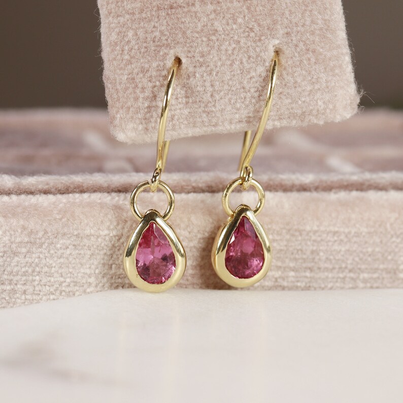 May include: A pair of gold teardrop earrings with pink gemstones. The earrings are hanging from a velvet box.