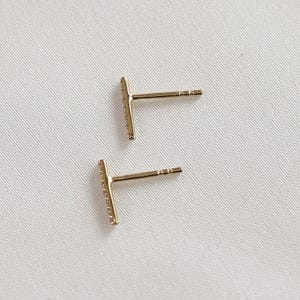 Back and side view of two gold diamond bar earrings showing the posts on a textured fabric.