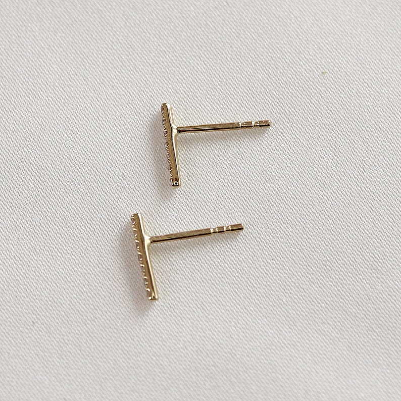 Back and side view of two gold diamond bar earrings showing the posts on a textured fabric.