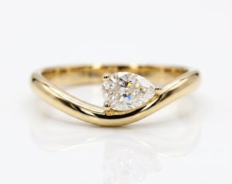 May include: A gold ring featuring a single pear-shaped clear gemstone set into a smooth, curved band. The metal has a polished finish, and the stone is positioned asymmetrically along the band, creating a sleek and continuous flowing design.