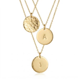 May include: Three gold-colored necklaces with circular pendants. One pendant has a hammered texture with a symbol, another has the letter "A", and the third has the letter "i". Each pendant hangs from a delicate chain.