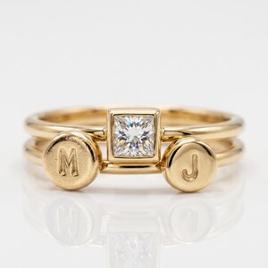Two stacked gold bands featuring a central square diamond ring flanked by two dangling circular initial charms stamped with M and J.