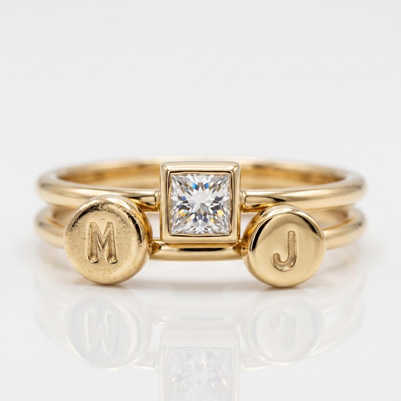 Two stacked gold bands featuring a central square diamond ring flanked by two dangling circular initial charms stamped with M and J.