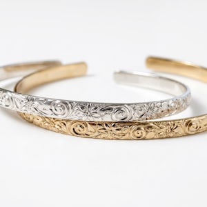 Three open cuff bracelets, two gold and one silver, with a floral and swirl pattern engraved on the outside.