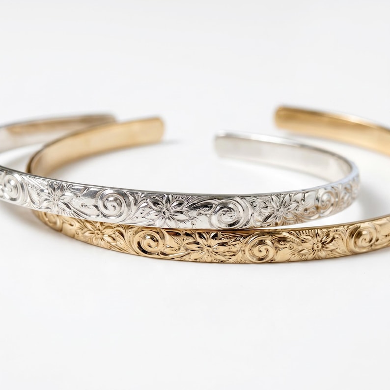 Three open cuff bracelets, two gold and one silver, with a floral and swirl pattern engraved on the outside.
