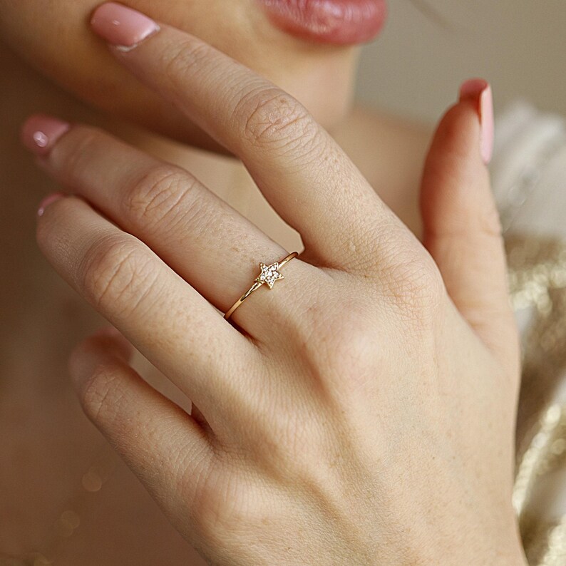 A woman's hand with light pink nail polish wears a thin gold ring with a small diamond-studded star on her ring finger.