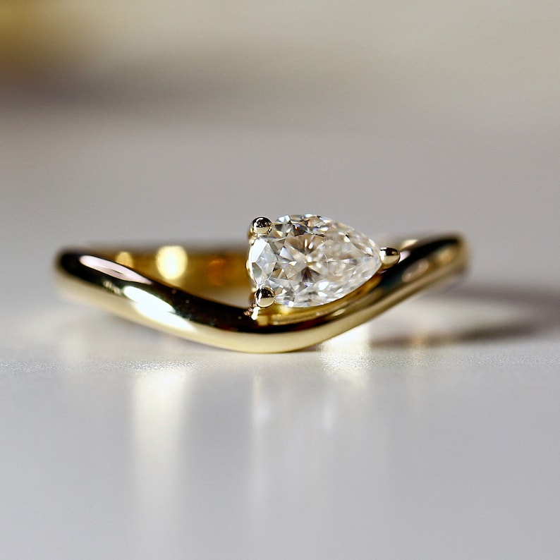 Pode incluir: Uma alian&ccedil;a de noivado de ouro com um diamante em forma de p&ecirc;ra engastado em uma banda curva.