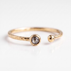May include: A gold open-style ring with a hammered texture on the band. One end of the ring features a round, bezel-set grey diamond, while the other end terminates in a small, smooth gold sphere. The metal has a warm, polished yellow finish.