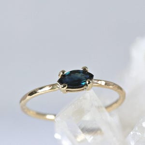 A gold ring with a dark blue marquise-cut sapphire set horizontally in four prongs, resting on a white crystalline surface.