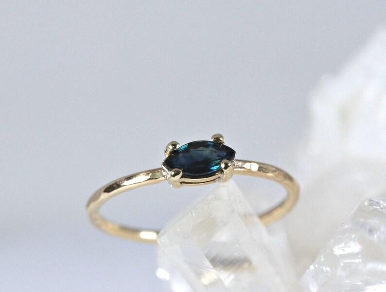 A gold ring with a dark blue marquise-cut sapphire set horizontally in four prongs, resting on a white crystalline surface.
