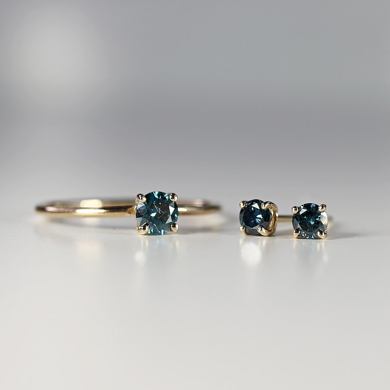 Puede incluir: Un conjunto de joyas de oro que incluye un anillo y un par de pendientes con piedras preciosas azules. El anillo tiene una sola piedra preciosa redonda azul engastada en una banda de oro. Los pendientes tienen una sola piedra preciosa redonda azul engastada en un engaste de oro.