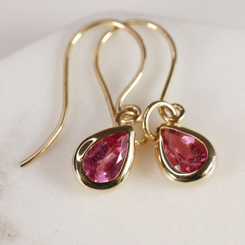May include: A pair of gold dangle earrings with pink teardrop-shaped gemstones.