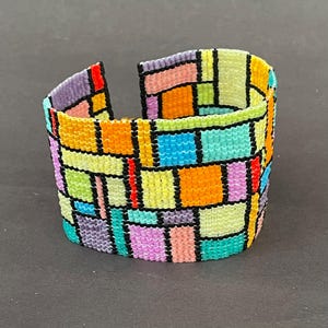 May include: A wide, open cuff bracelet with a colorful, geometric design. The bracelet is made of small, woven beads in shades of orange, yellow, green, blue, purple, and red, outlined with black. The bracelet is approximately 2 inches wide.