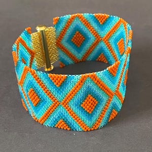 May include: A beaded cuff bracelet featuring a geometric diamond pattern in shades of turquoise and orange. The bracelet has a gold-colored clasp. The design is a repeating pattern of diamonds.