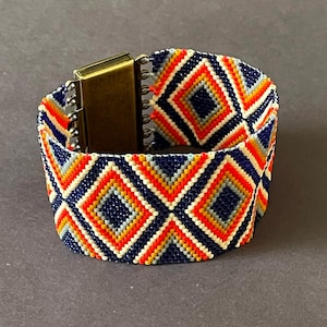May include: A wide beaded bracelet with a geometric diamond pattern in navy blue, orange, yellow, and white. The bracelet has a bronze-colored magnetic clasp.