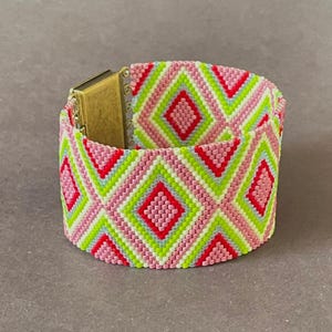 May include: A wide beaded bracelet with a geometric pattern of pink, red, green, and white diamonds. The bracelet has a gold-colored clasp. The bracelet is approximately 2 inches wide.