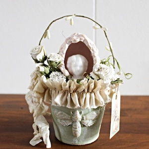 May include: A decorative, light green pot with a moth design, filled with white roses and greenery. A small, white-faced figurine is nestled within the arrangement, under a draped, ruffled fabric. A tag hangs from the pot.