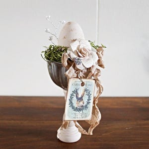 May include: An Easter-themed decorative egg cup. The egg cup is made of metal and has a white base. It is decorated with an egg, flowers, greenery, and a tag with a rabbit illustration. The color palette includes beige, white, and green.