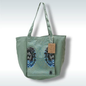 Pode incluir: Bolsa tote verde sálvia com um gráfico de leão com detalhes em azul e preto. A bolsa tem alças longas e um pequeno bolso com o desenho do leão. Uma etiqueta castanha pende da alça.