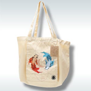 Pode incluir: Bolsa tote creme com um bolso com um desenho de dois peixes koi, um vermelho e outro azul, nadando em torno de uma lua crescente. A bolsa tem alças longas e uma pequena etiqueta.