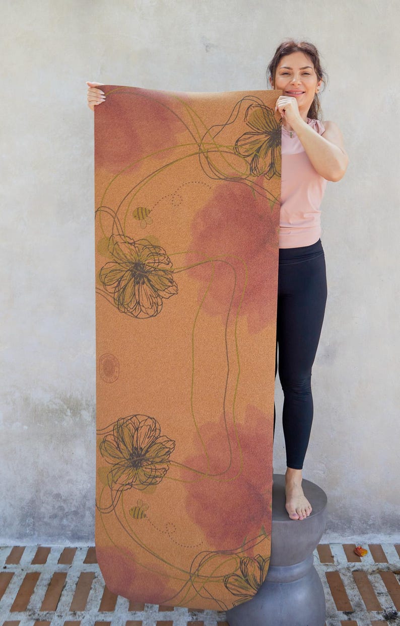 May include: A cork yoga mat with a floral design in shades of brown, green, and pink. The mat features a repeating pattern of stylised flowers and bees.
