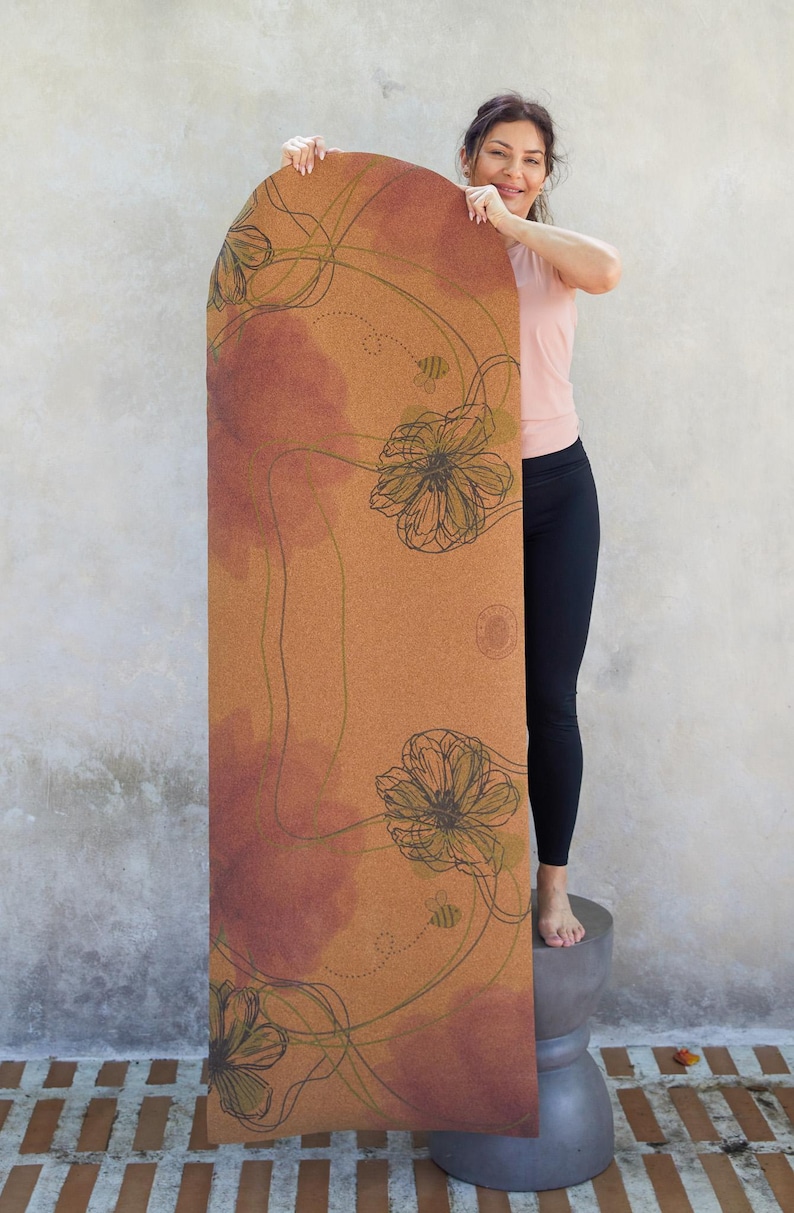 May include: A cork yoga mat with a floral design in shades of brown, green, and pink. The mat features a repeating pattern of stylised flowers and bees.