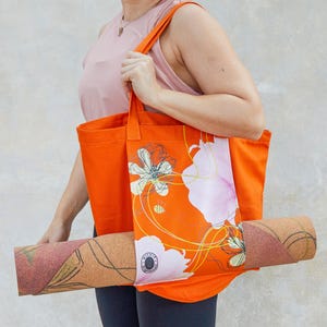 May include: A bright orange tote bag with a floral design and a yoga mat tucked inside. The bag has a white floral pattern with yellow lines and a bee. The yoga mat is brown with a cork texture.