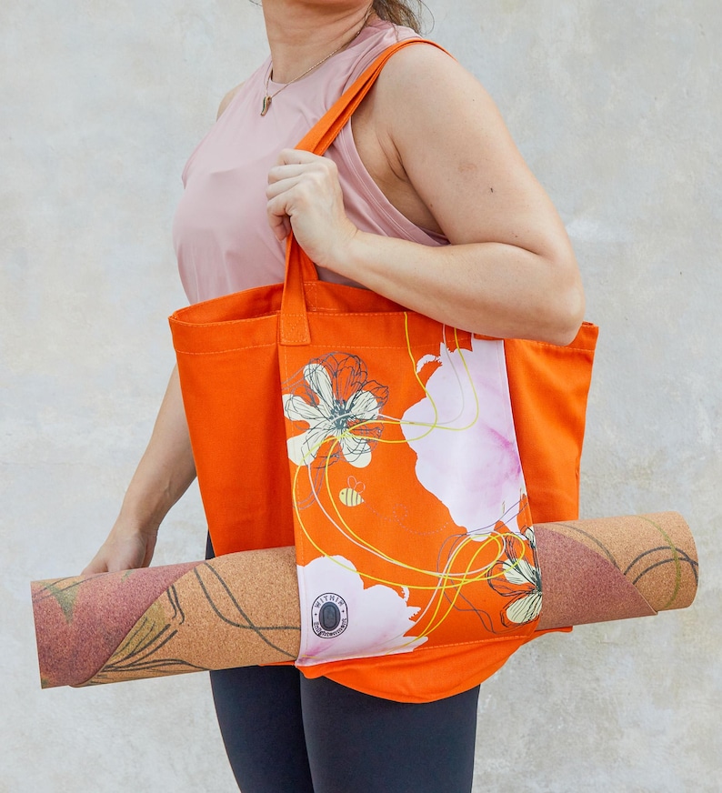 May include: A bright orange tote bag with a floral design and a yoga mat tucked inside. The bag has a white floral pattern with yellow lines and a bee. The yoga mat is brown with a cork texture.