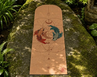 May include: A rectangular tan yoga mat featuring a central graphic of two koi fish, one orange and one teal, swimming in a circle. The design includes thin brown geometric lines, celestial moon phases, and a small circular logo in the bottom corner.