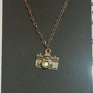 Könnte beinhalten: Eine silberne Kettenkette mit einem silbernen Kamera-Anhänger. Der Kamera-Anhänger hat ein detailliertes Design mit einem Objektiv und einer strukturierten Oberfläche.