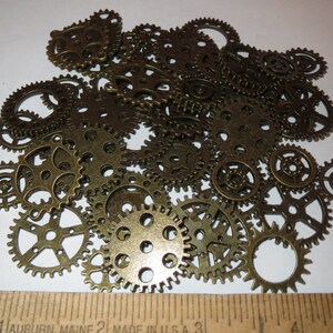 May include: A collection of antique gold-toned metal gears of various sizes. The gears are arranged in a random pattern on a white surface.